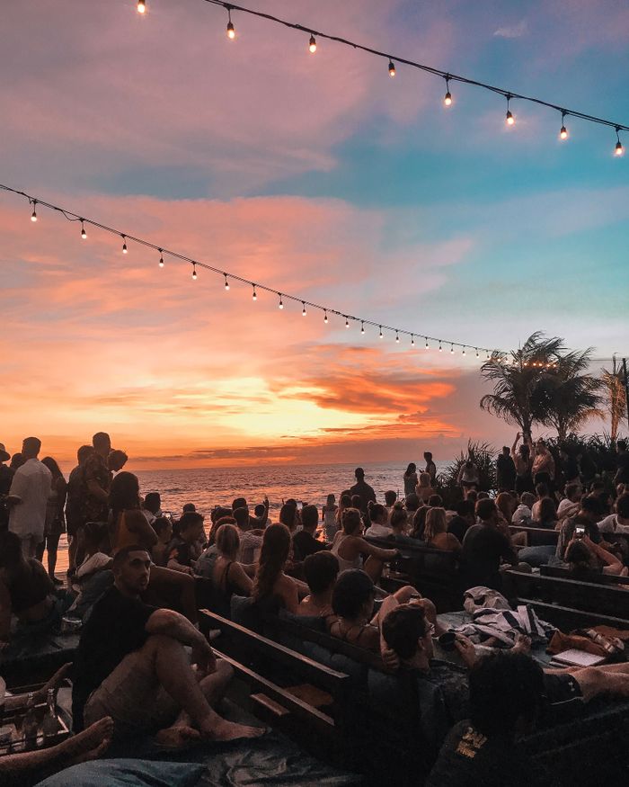 Crowd enjoying a stunning sunset at a Bali beach with string lights and palm trees.
