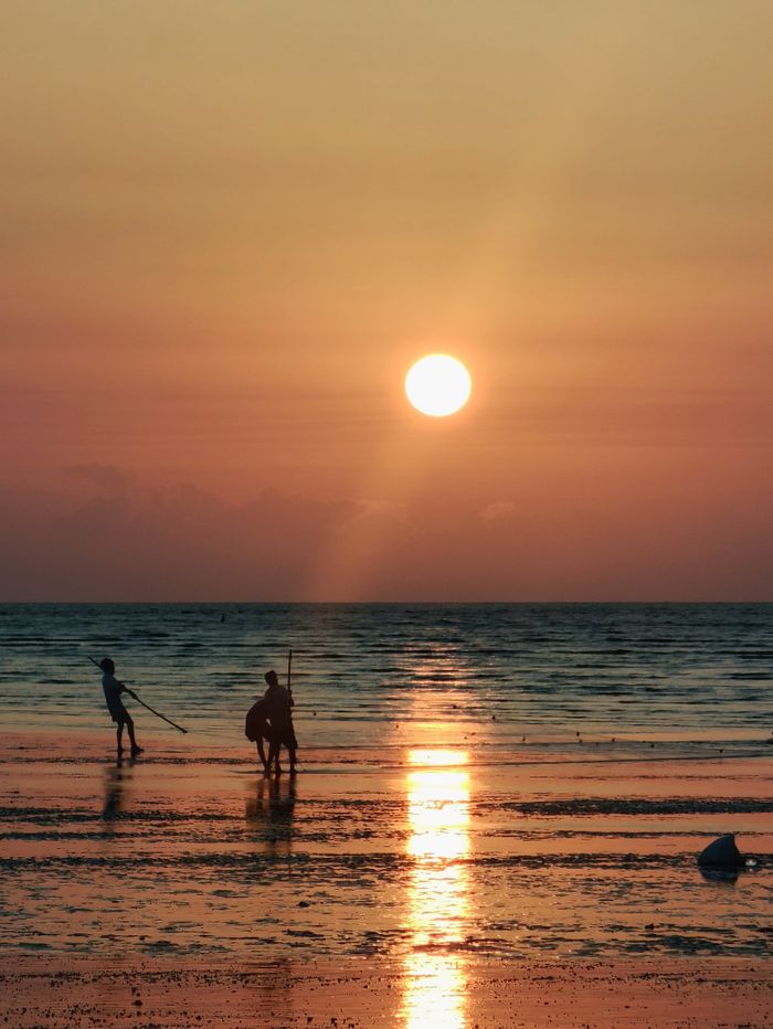 Two fishermen silhouetted against a vibrant sunset on a calm beach, showcasing serene coastal life.