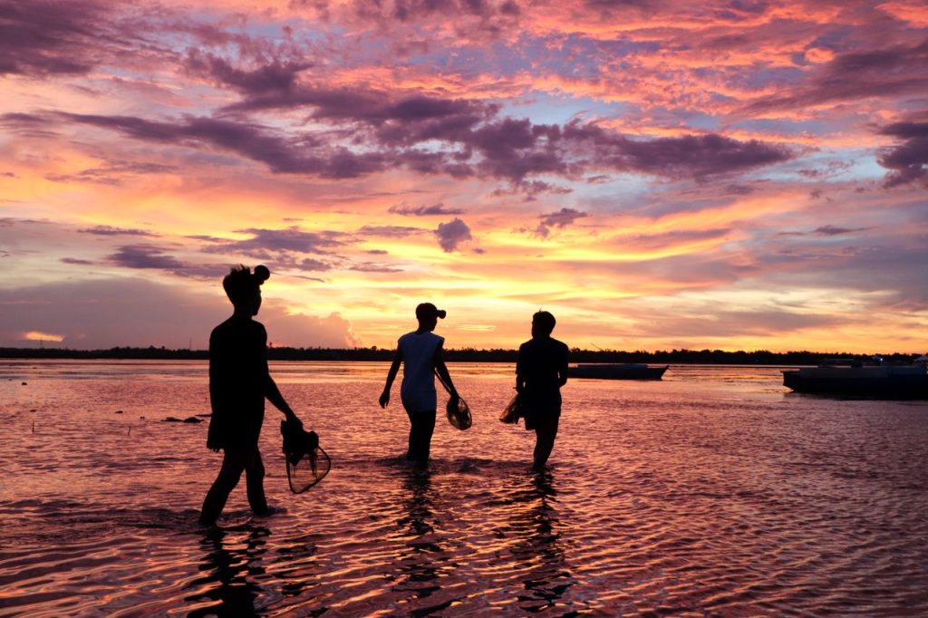 Three silhouetted fishermen walking through shallow waters at sunset in Bogo City, Philippines.