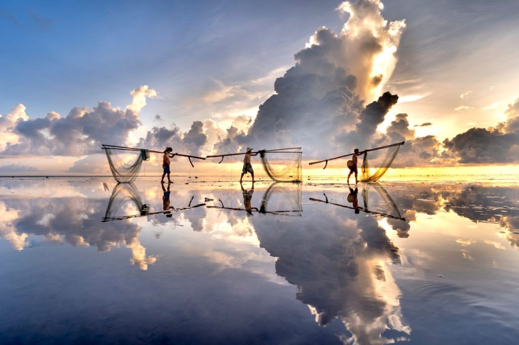 Three fishermen carrying nets reflect against calm water at sunset, creating a stunning silhouette scene.